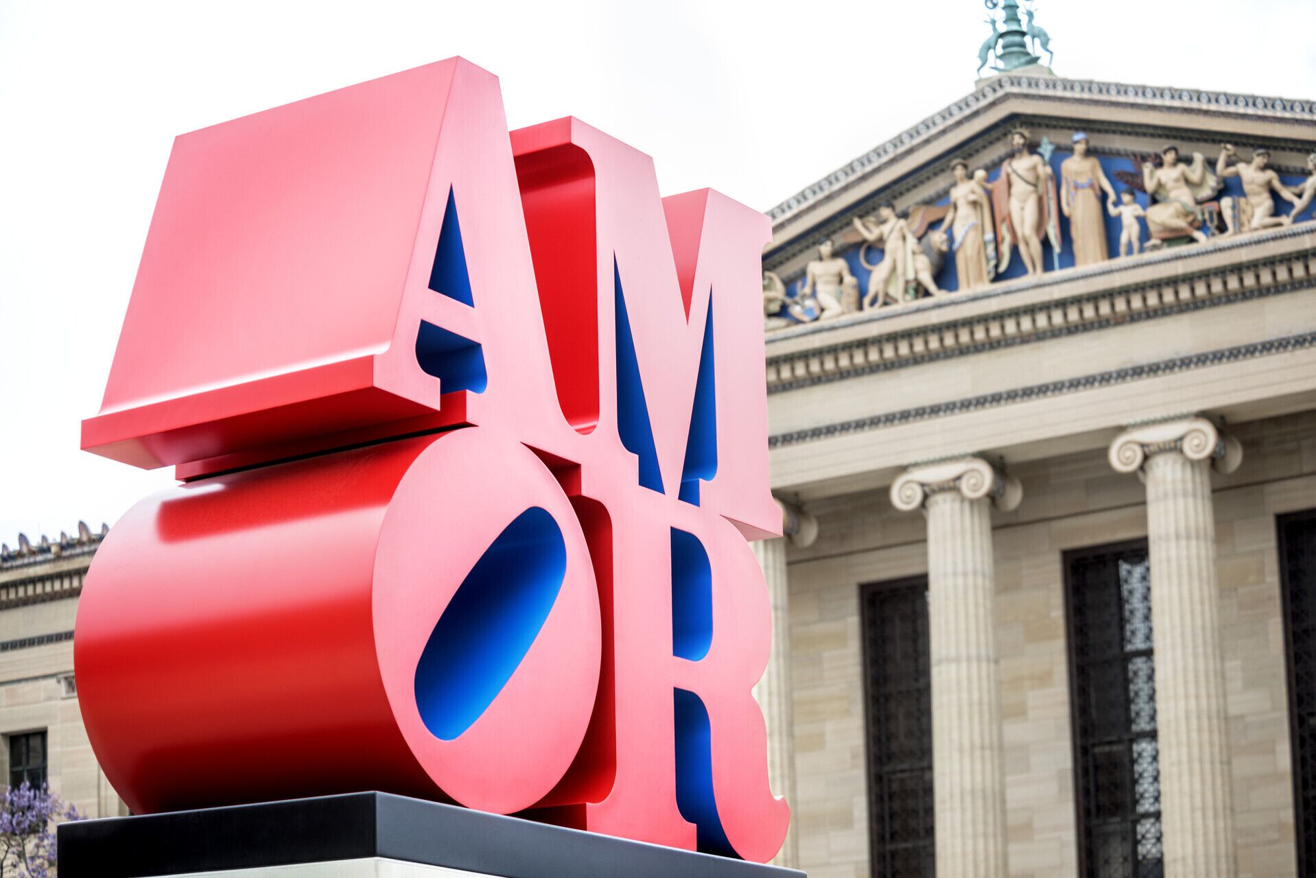 Una gran escultura roja y azul con las letras “A” y “V” bien visibles frente a un edificio clásico con columnas y estatuas.