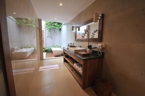 Un baño moderno con una bañera grande, un lavamanos y un espejo. Se aprecia un estante con toallas y un cesto de basura.