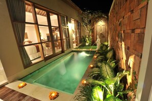 A swimming pool with a wooden deck, surrounded by a brick wall and lush greenery. - Maitri Villa (Ubud)