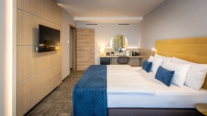 A hotel room with a large bed, a desk with a chair, a television mounted on the wall, and a small kitchenette area. - Nádas Tó Park Hotel (Vasad)
