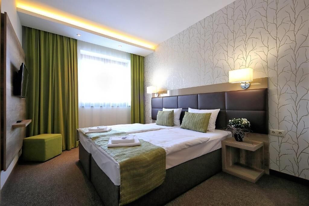 Una habitación de hotel con una cama grande, una mesita de noche con lámpara, un televisor montado en la pared y una ventana con cortinas.