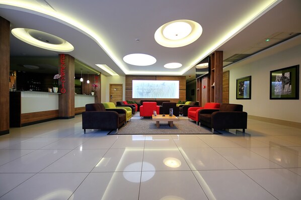 Un hall d'entrée moderne avec un comptoir d'accueil, des sièges aux couleurs vives et un grand écran diffusant une présentation.