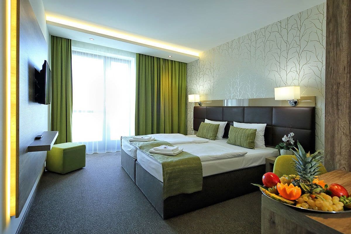 Ein Hotelzimmer mit einem großen Bett, zwei Nachttischen, einem grünen Hocker, einer Obstschale und einem an der Wand befestigten Fernseher.