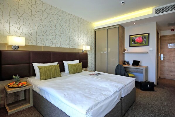 A hotel room with a large bed, two bedside lamps, a wooden nightstand with fruit, a desk with a laptop, and a sliding door closet. - Nádas Tó Park Hotel (Vasad)