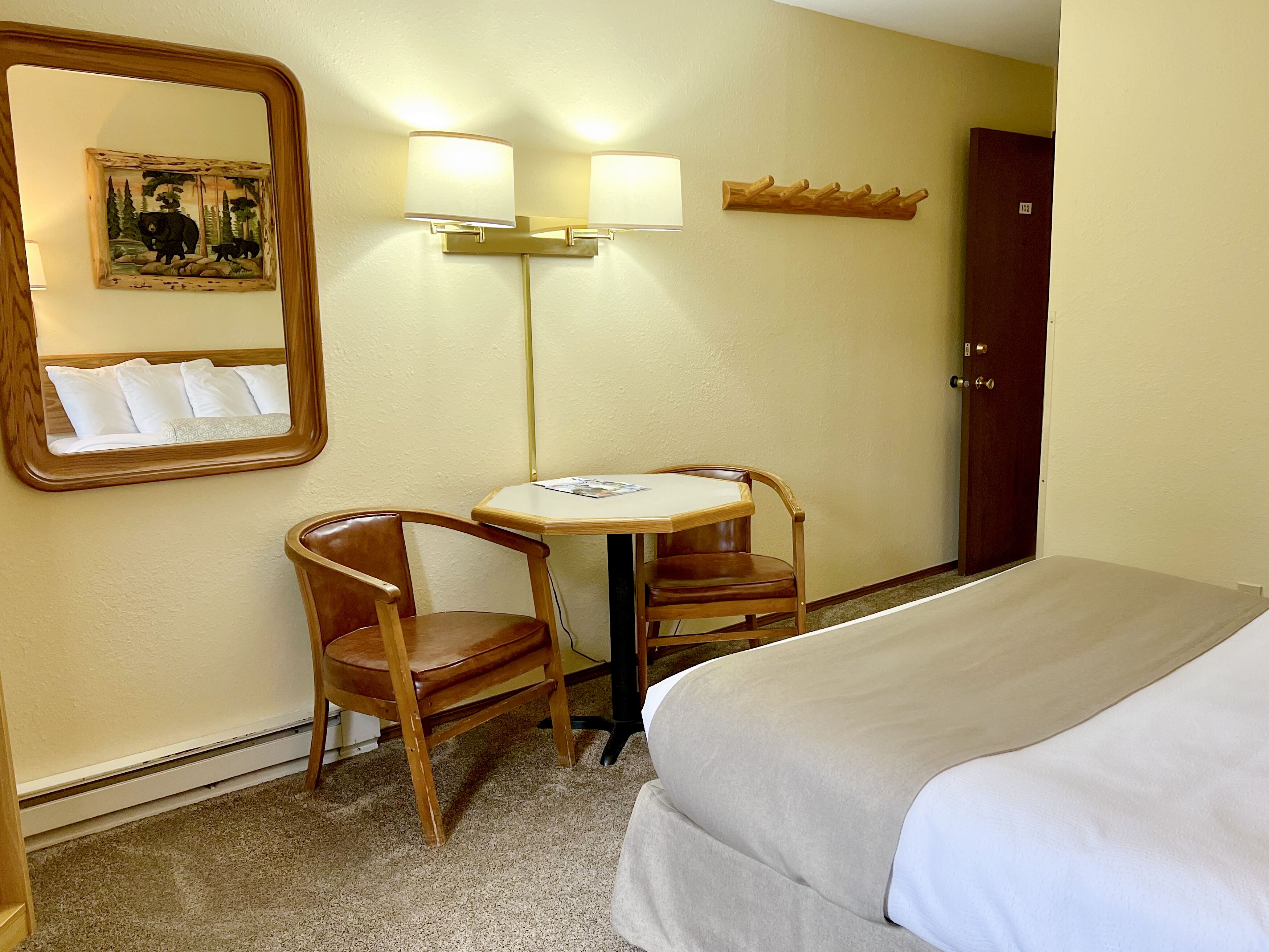 A hotel room with a bed, two wooden chairs, a small table, a mirror, and a wall-mounted lamp.