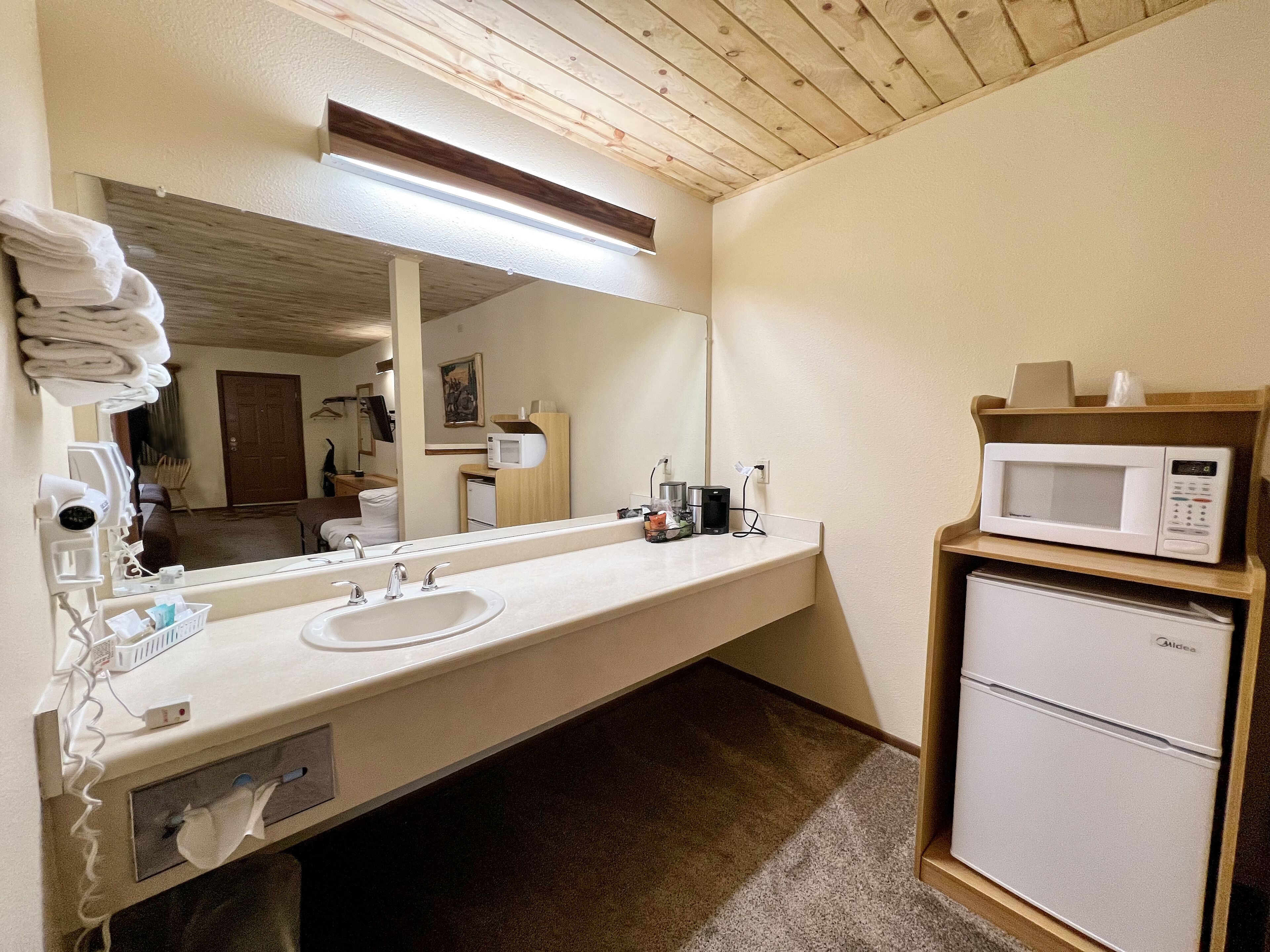 A hotel bathroom with a sink, mirror, towel rack, and a microwave on a shelf.