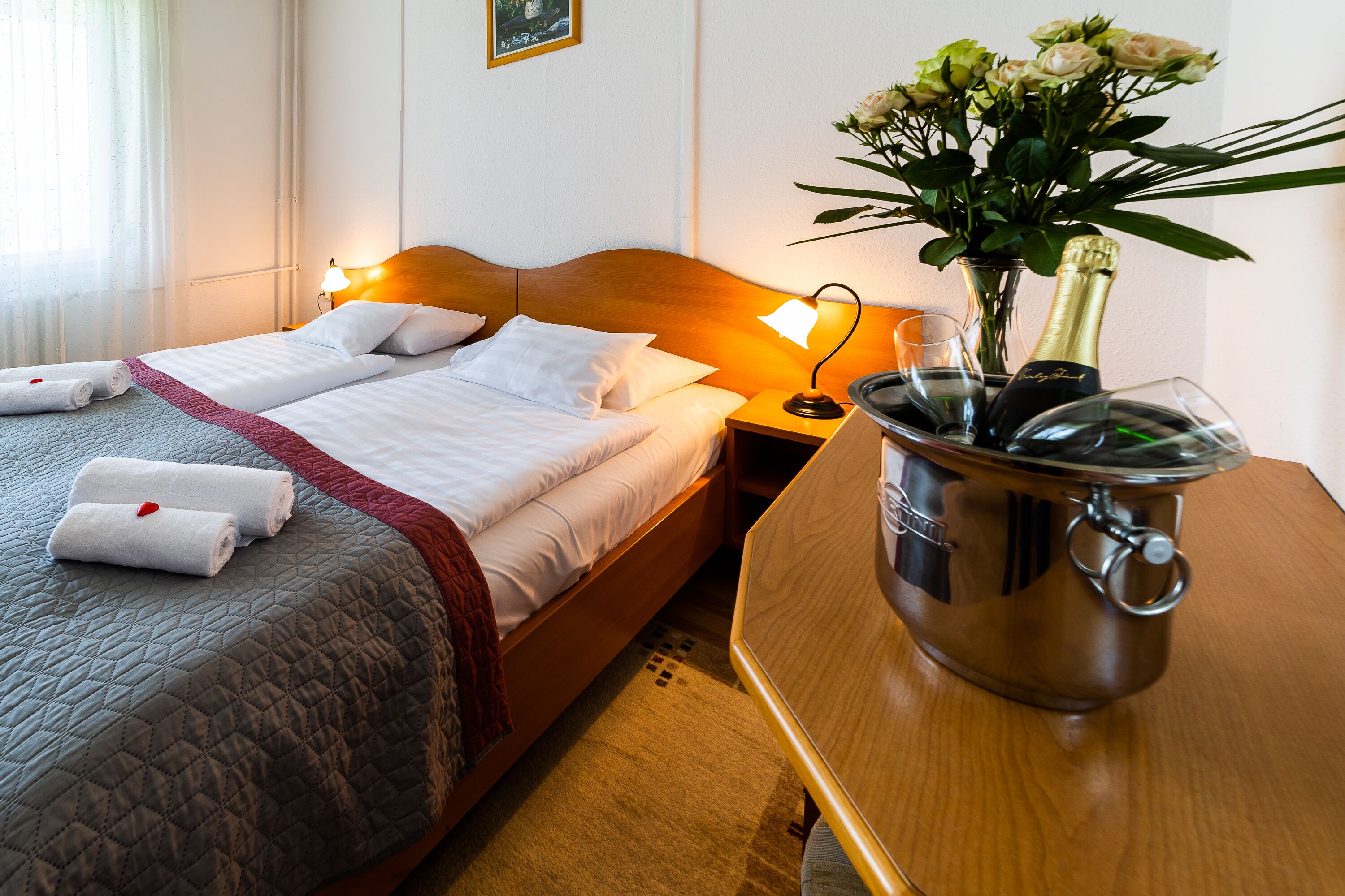 A hotel room with a bed, bedside table, and a nightstand with a lamp. There is a vase with flowers on the nightstand.