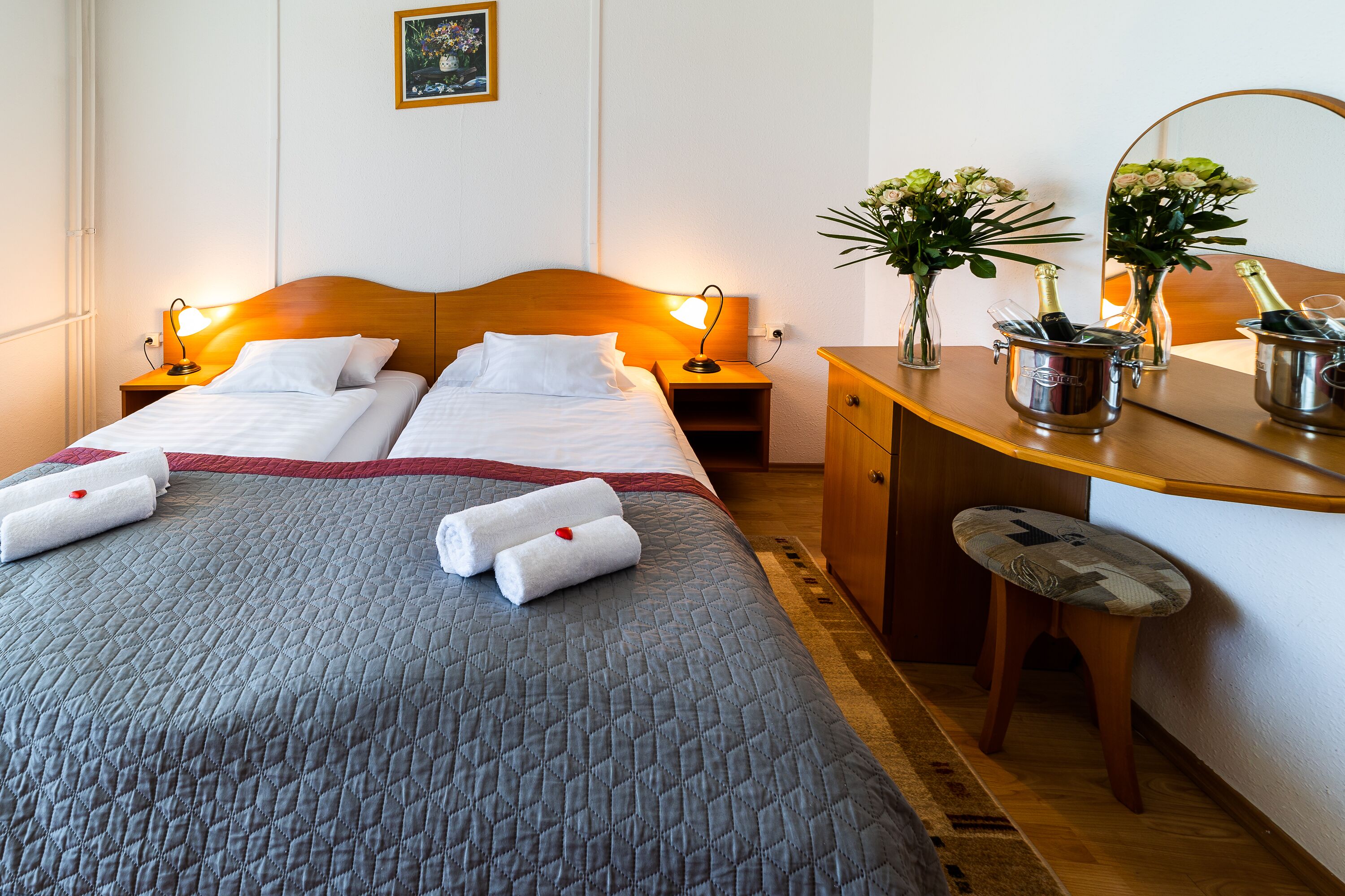 Habitación de hotel con dos camas, un cabecero de madera, lámparas de noche, un escritorio de madera con un espejo, un taburete pequeño y un jarrón con flores.