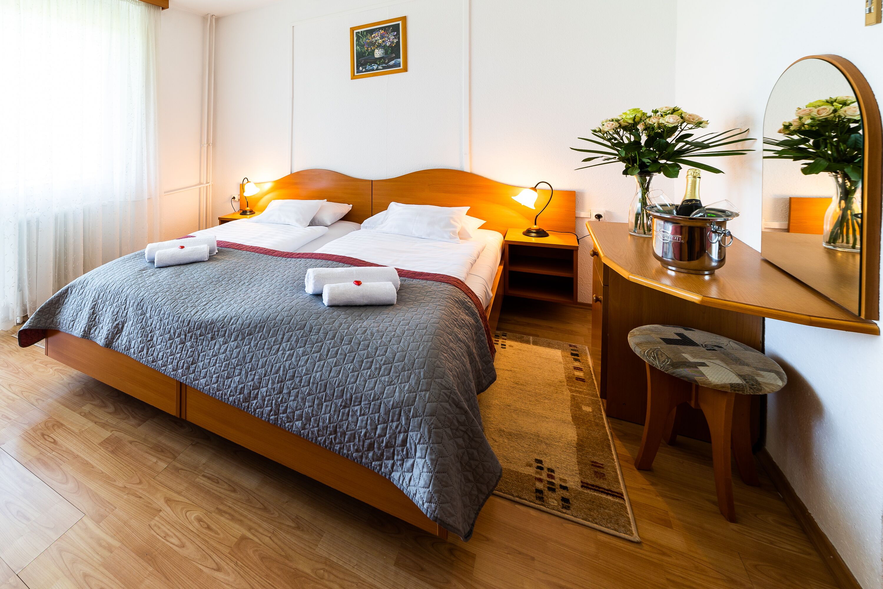 Habitación de hotel con una cama de madera, una mesita de noche con lámpara, un escritorio con espejo y una planta en maceta.