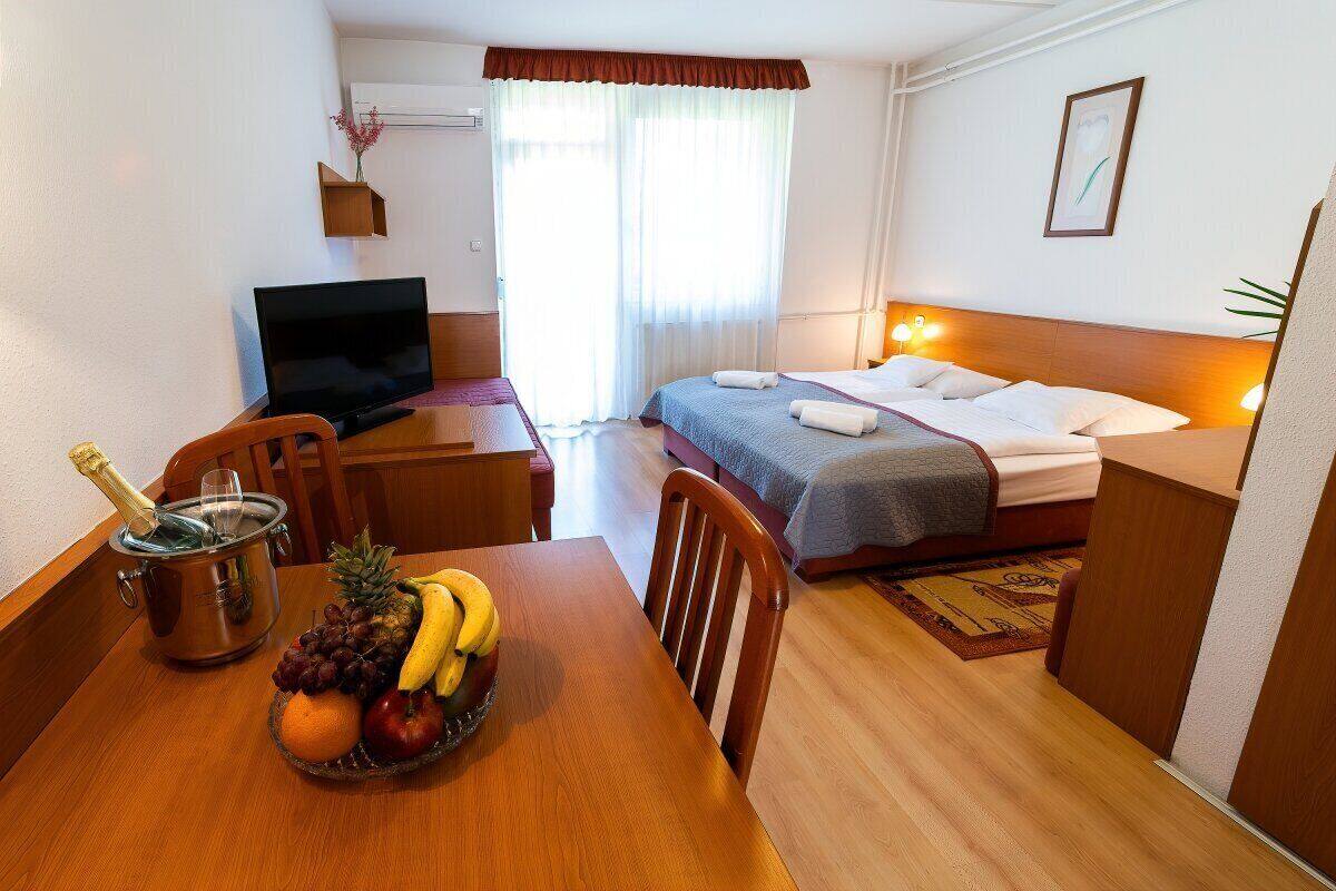 A hotel room with a bed, a TV, a dining table with fruit and a champagne bottle, and a window with curtains.