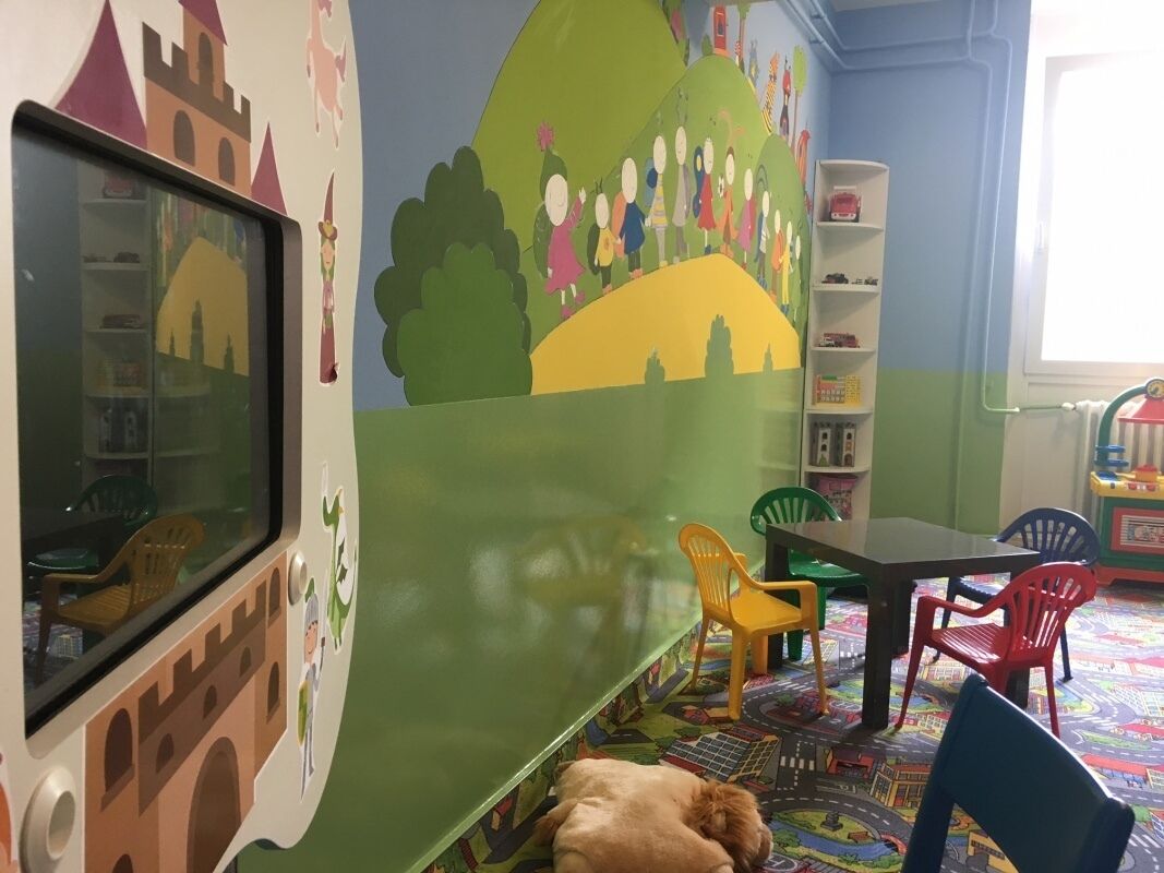 Ein Zimmer mit großem Fenster, einem Bücherregal, einem Schreibtisch und einer bunten Spielmatte.