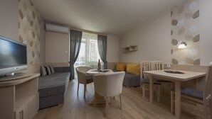 A modern living room with a sofa, dining table, chairs, a TV, and a window with curtains. - Hotel Napfény (Zalakaros)