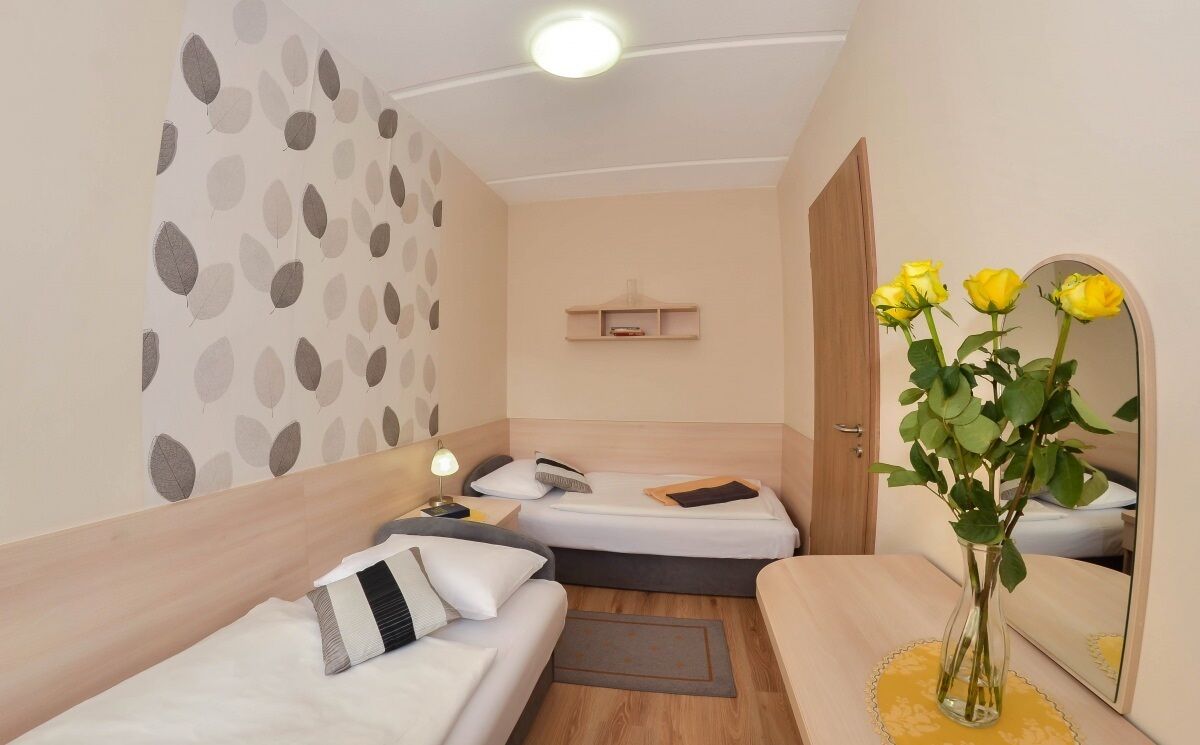 A small, cozy room with two beds, a bedside table with a vase of flowers, a mirror, and a shelf with books.