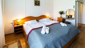 A neatly made double bed with a blue quilt, two bedside lamps, a nightstand, a mirror, and a small table with a vase of flowers. - Hotel Napfény (Zalakaros)