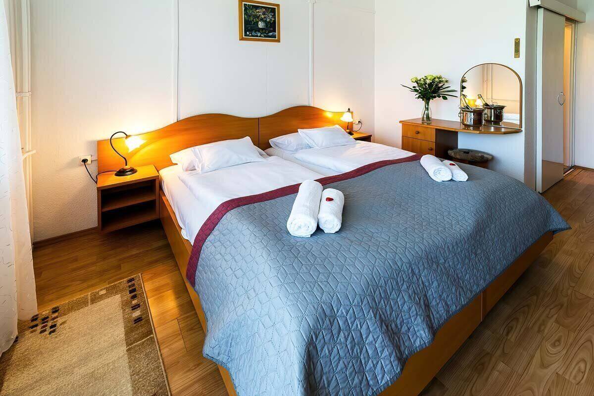 A neatly made double bed with a blue quilt, two bedside lamps, a nightstand, a mirror, and a small table with a vase of flowers.