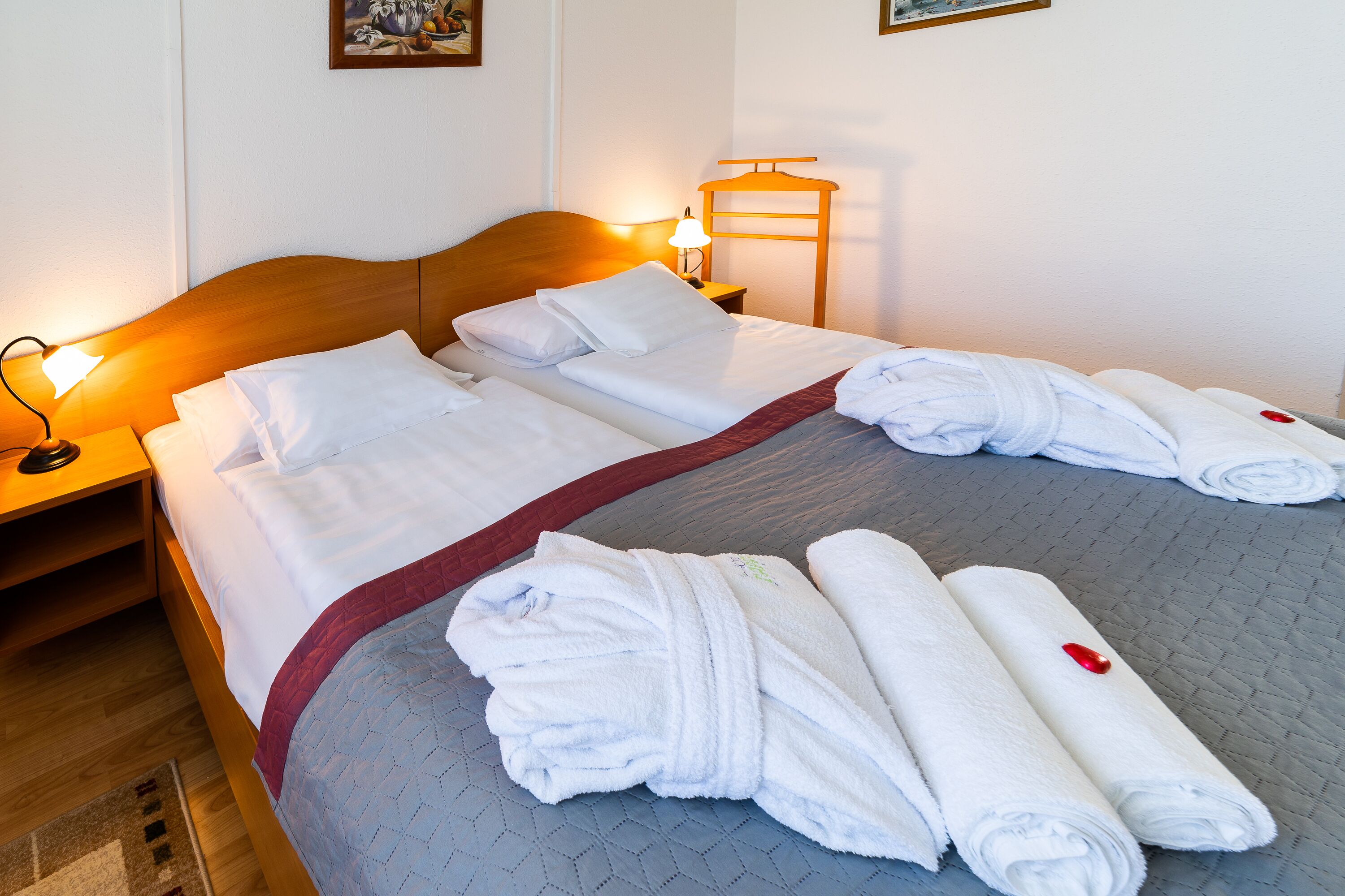 A neatly made bed with white linens and rolled towels, a wooden headboard, and a bedside table with a lamp.