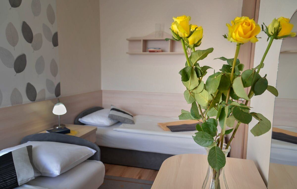 Habitación de hotel con cama, mesita de noche con lámpara, una pequeña estantería y un jarrón con rosas amarillas.