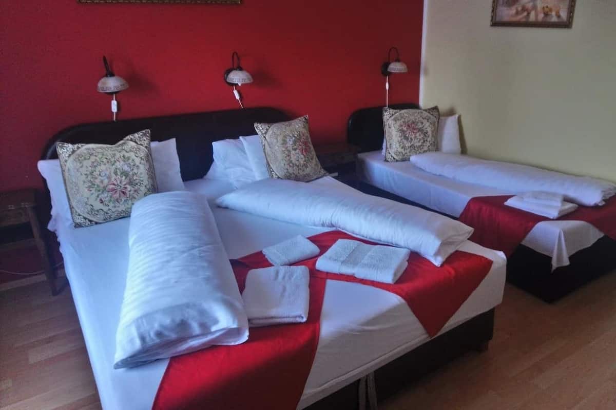 A hotel room with three beds, each with white linens and red bedspreads. There are wall-mounted lamps on the left side of the beds.