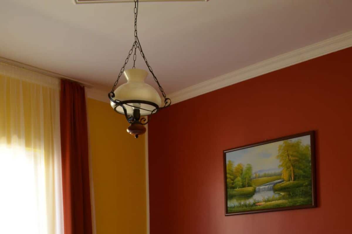 A neatly made bed with white and orange bedding, a painting of a landscape on the wall, and a hanging light fixture.