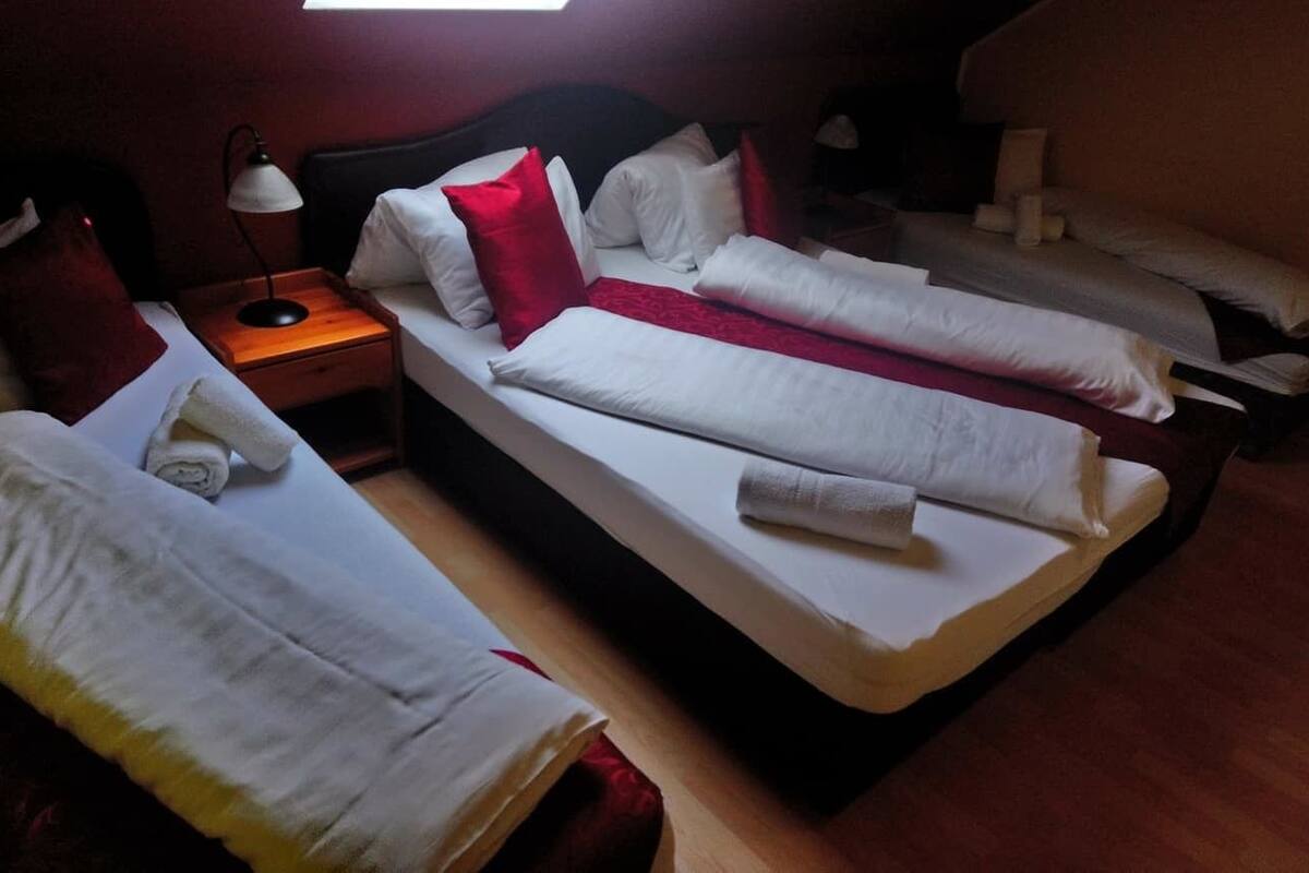 A hotel room with three beds, a nightstand with a lamp, and a window providing natural light.