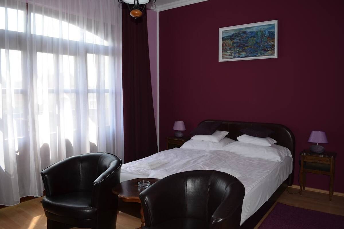 A hotel room with a bed, two nightstands, a painting on the wall, and a window with sheer curtains.