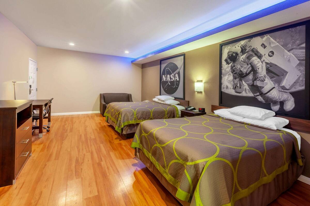 A hotel room with two beds, wooden flooring, a desk with a chair, a nightstand, and a NASA astronaut poster.