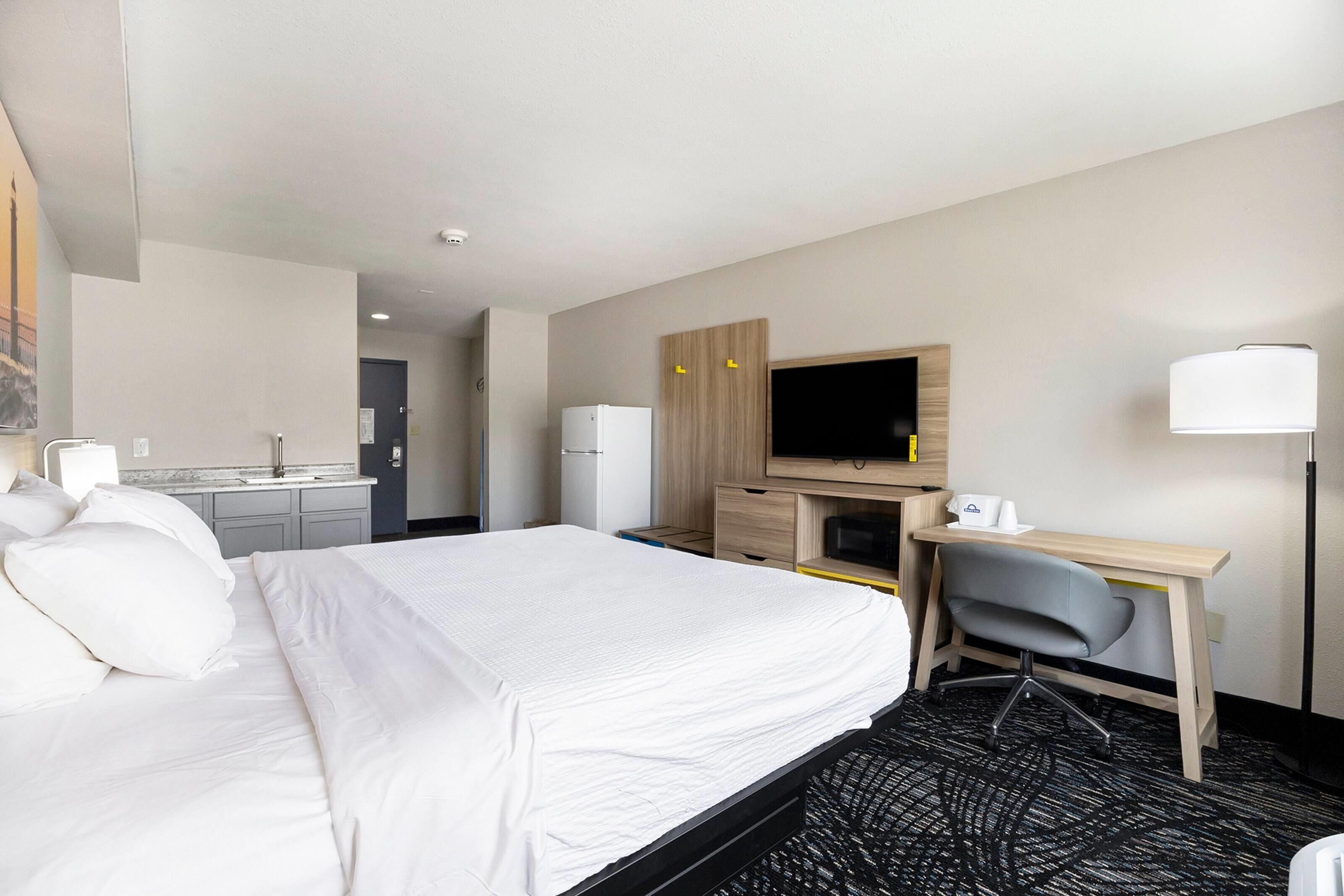 A hotel room with a bed, desk, chair, television, and kitchenette.