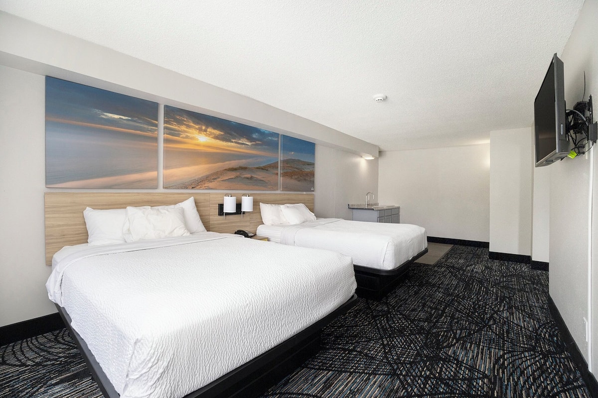 Habitación de hotel con dos camas, un gran mural en la pared con un atardecer desértico, un televisor de pantalla plana montado en la pared y una alfombra estampada.