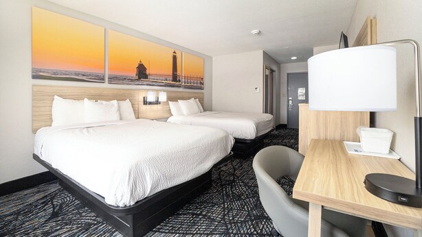 A hotel room with two beds, a desk with a lamp, a chair, and a painting of a lighthouse on the wall.