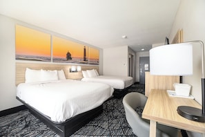 A hotel room with two beds, a desk with a lamp, a chair, and a painting of a lighthouse on the wall.