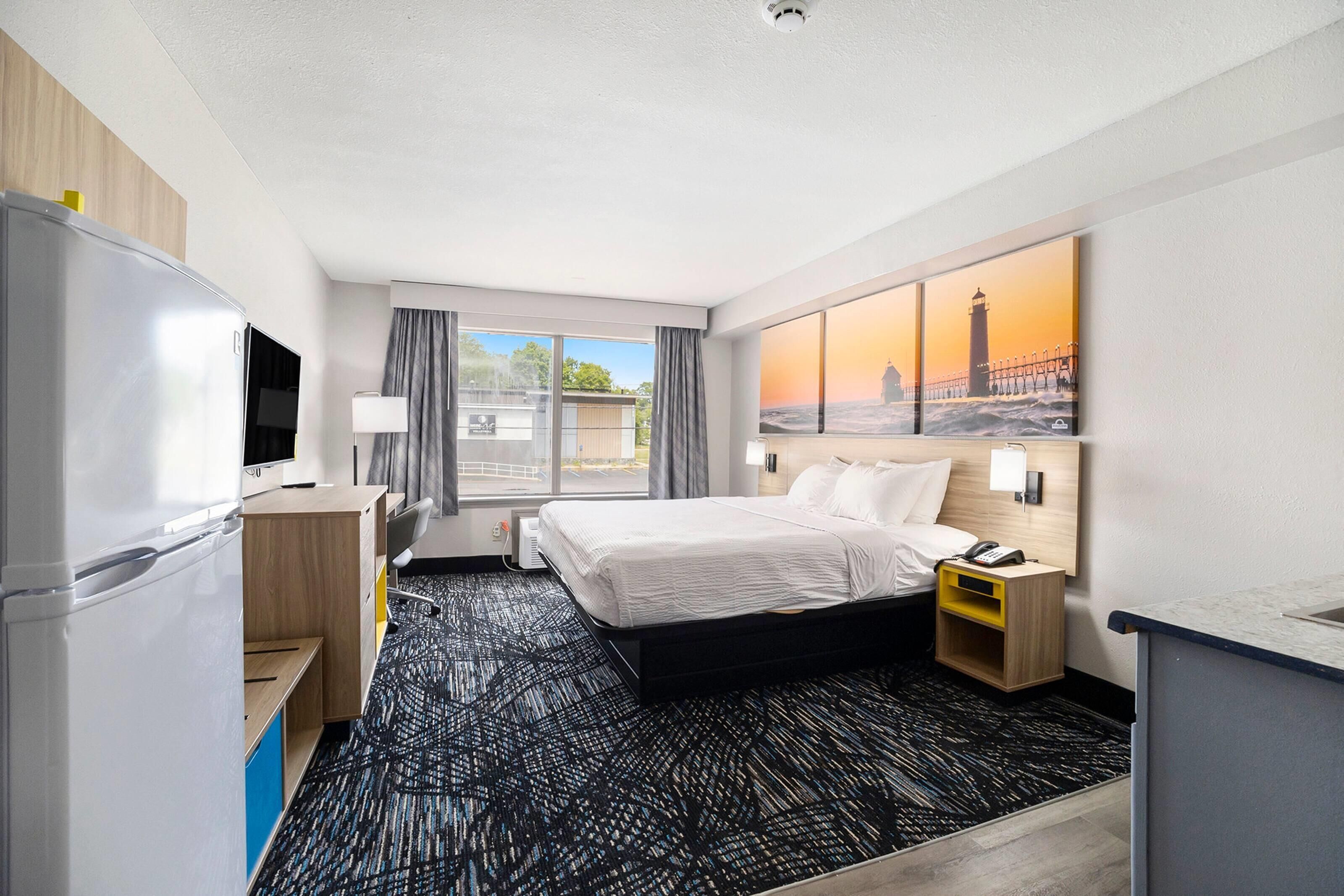 A hotel room with a bed, a desk, a refrigerator, a TV, a lamp, a nightstand, and a painting of a lighthouse.
