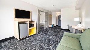 A hotel room with a flat-screen TV, a mini-fridge, a sofa, a desk with a lamp, and a patterned carpet.