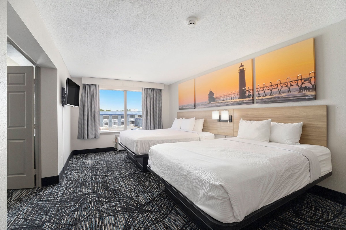 Habitación de hotel con dos camas, un ventanal con cortinas, un cuadro de un faro y una alfombra estampada.