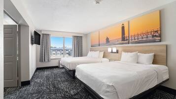 Habitación de hotel con dos camas, un ventanal con cortinas, un cuadro de un faro y una alfombra estampada.