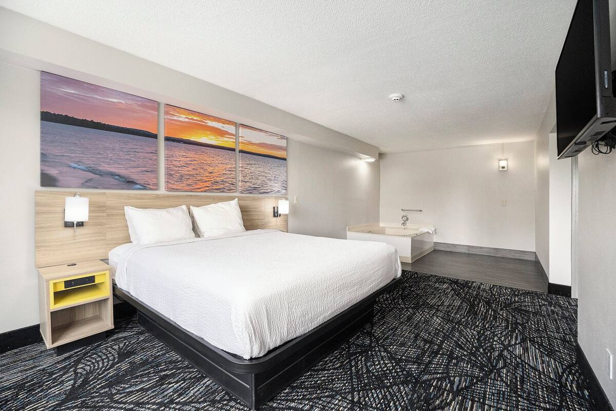 Habitación de hotel con una cama grande, una mesita de noche, un atril, un televisor montado en la pared y dos cuadros de un atardecer sobre el agua.