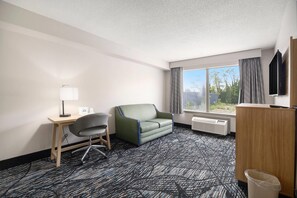 A hotel room with a desk, chair, sofa, TV, and a window with curtains. - Days Inn & Suites by Wyndham Norton Shores/Muskegon (Muskegon)