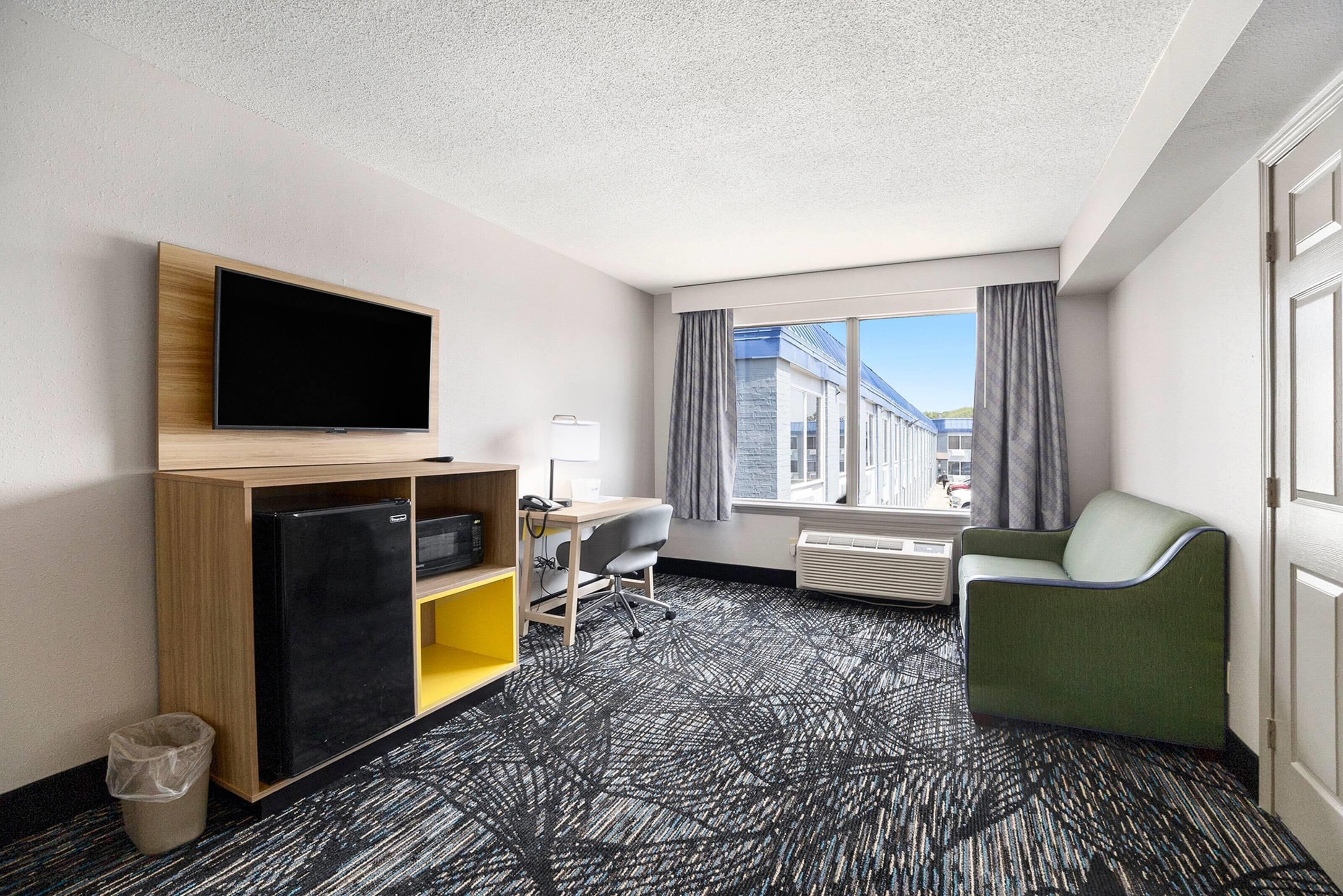 A hotel room with a flat-screen TV, a desk with a chair, a sofa, a waste bin, and a large window with curtains.