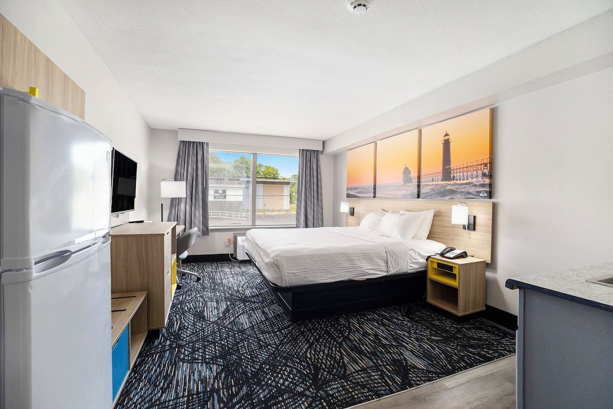 Habitación de hotel con cama, escritorio, refrigerador, televisor, ventana con cortinas y un cuadro de un faro.