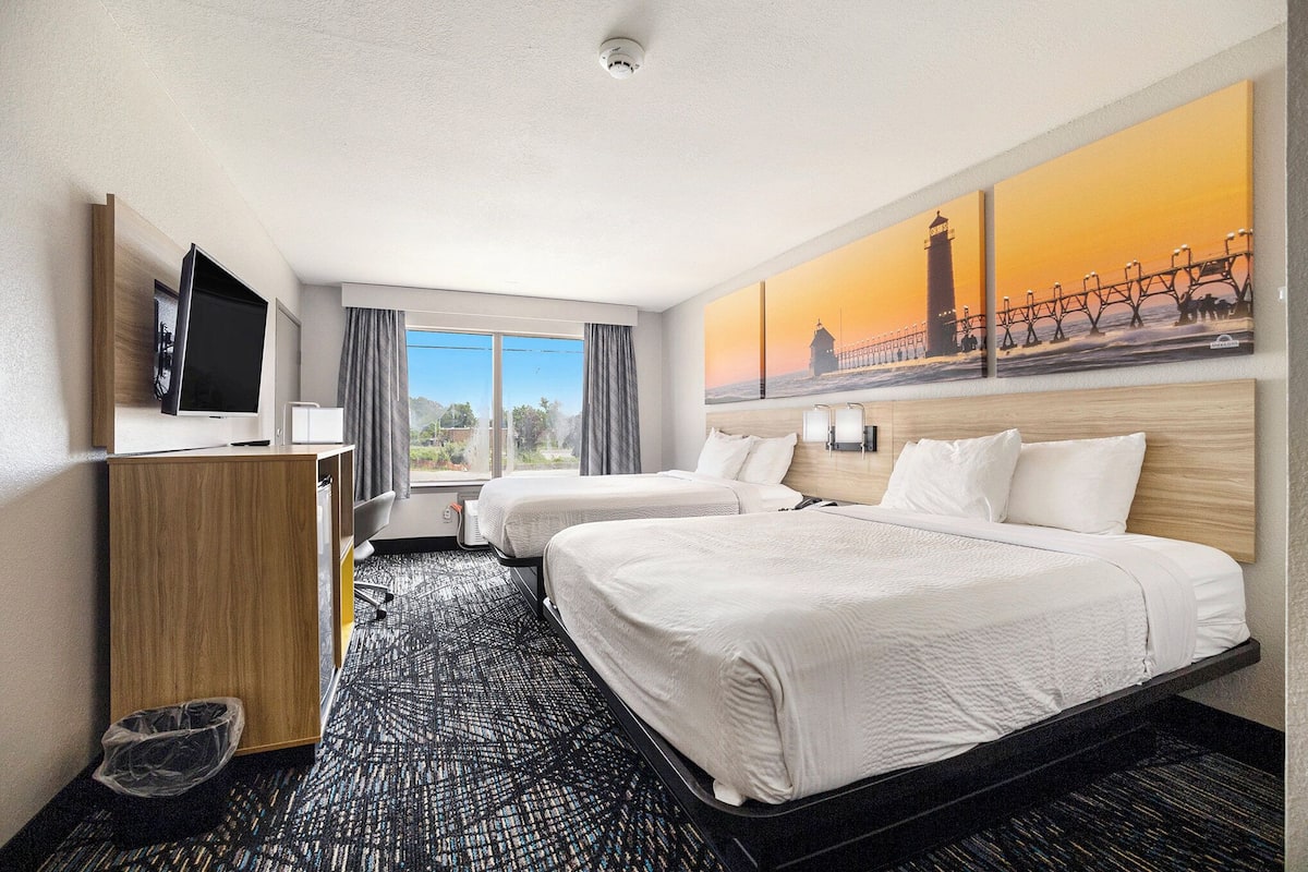 Habitación de hotel con dos camas, televisión, escritorio y ventana grande con vistas a un puente y un faro.