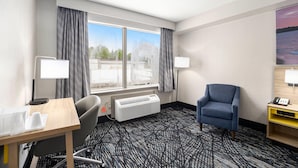 A hotel room with a large window, a desk with a chair, a blue armchair, a heater, and a lamp.