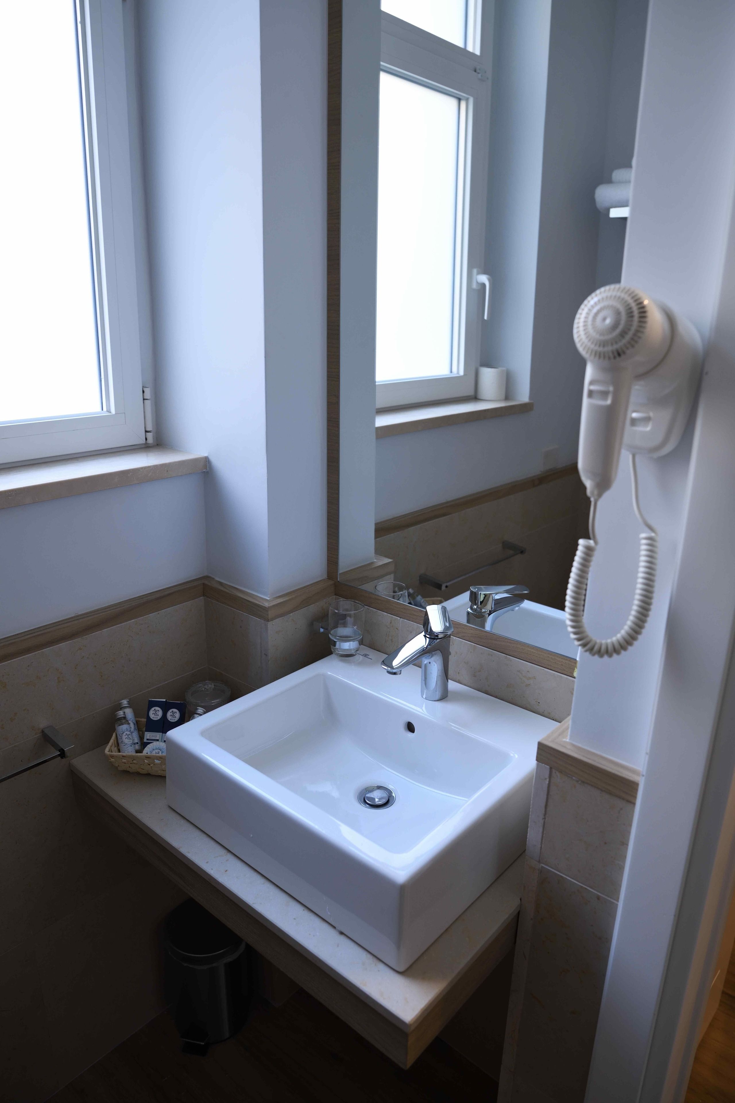 Ein Badezimmer mit einem weißen Waschbecken, einem Spiegel, einem Fenster und einem an der Wand befestigten Fön.
