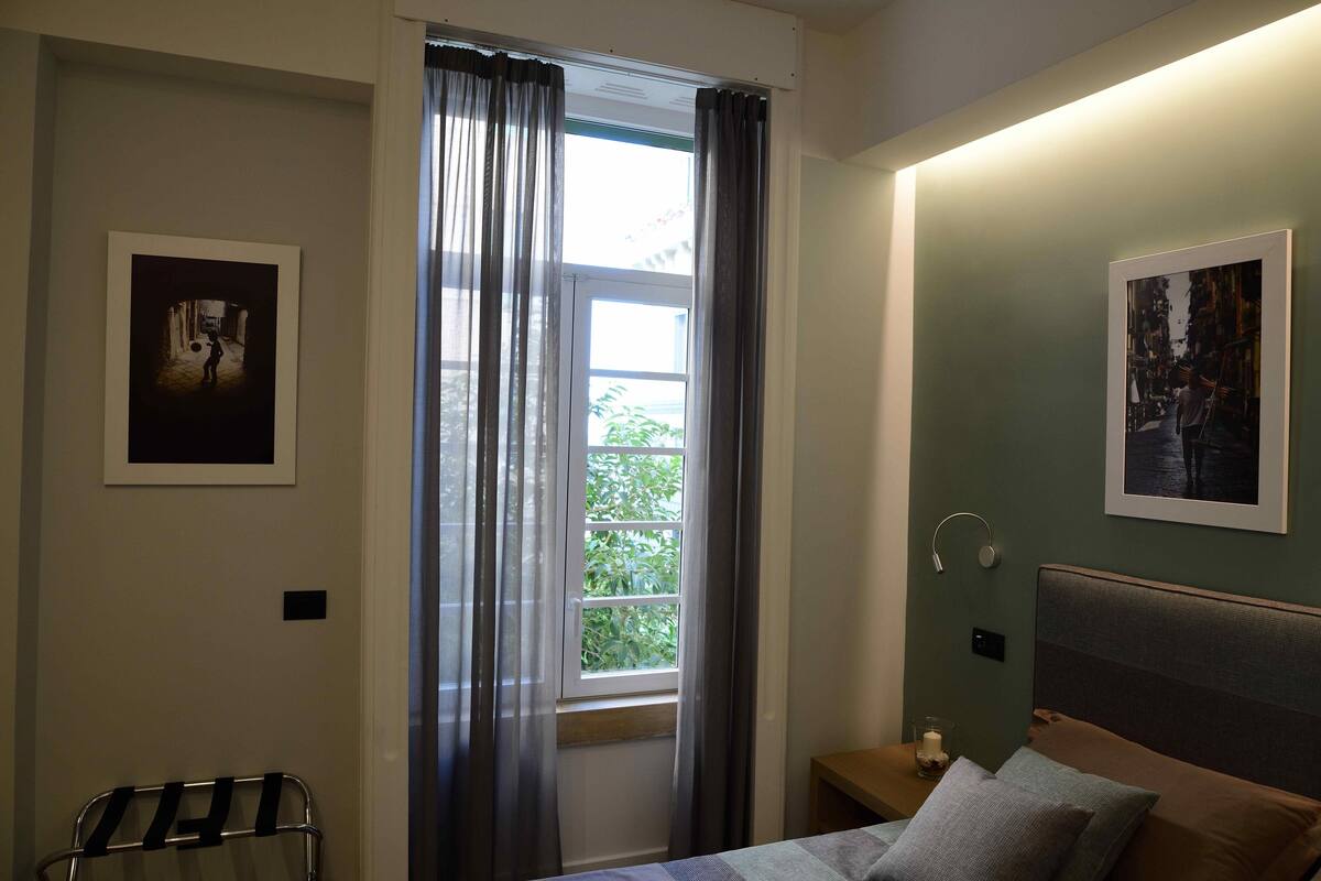 Quarto com cama, mesa de cabeceira, janela com cortinas e duas quadros na parede.