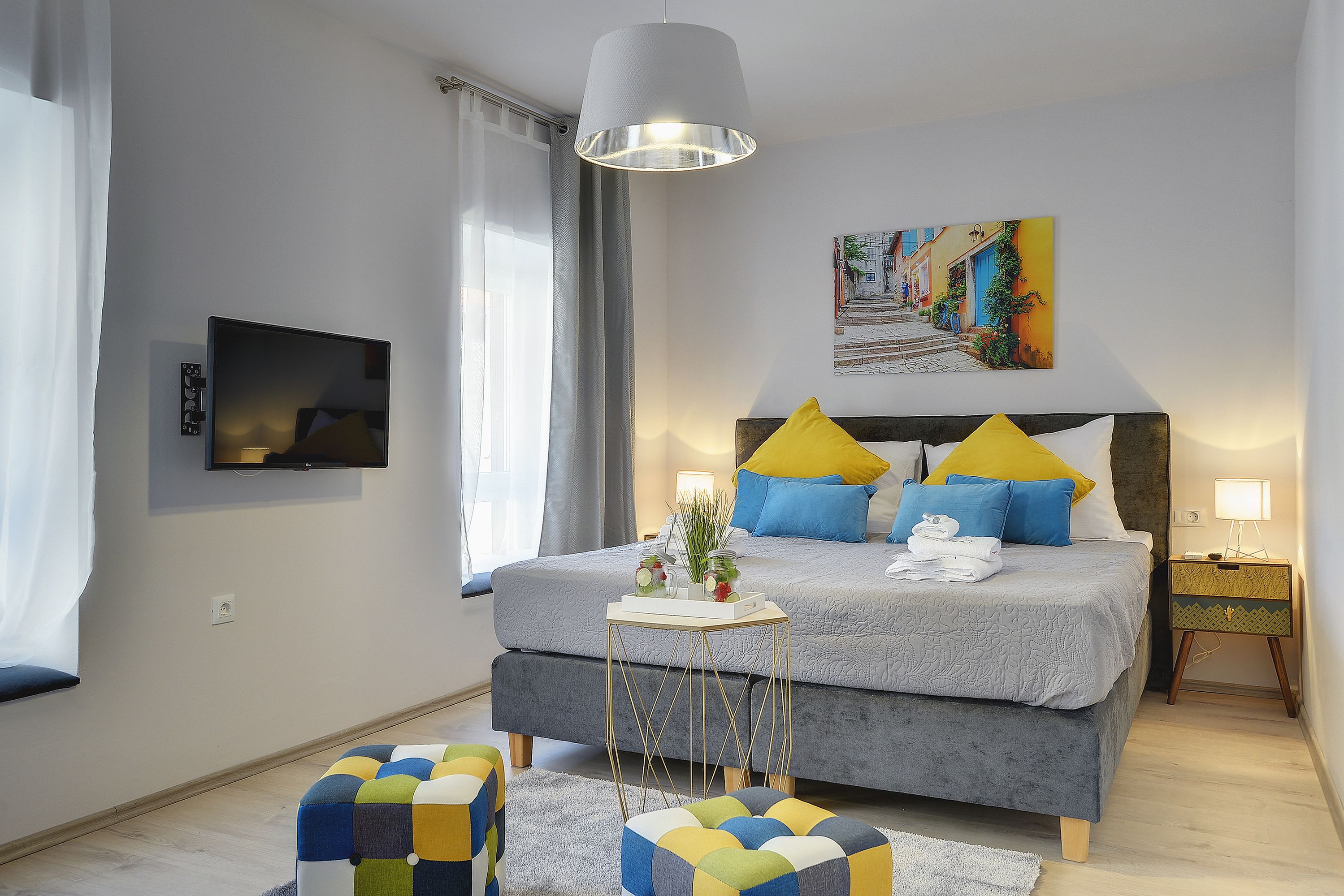 A modern bedroom with a grey bed, yellow and blue pillows, a small bedside table with a lamp, and a painting on the wall.
