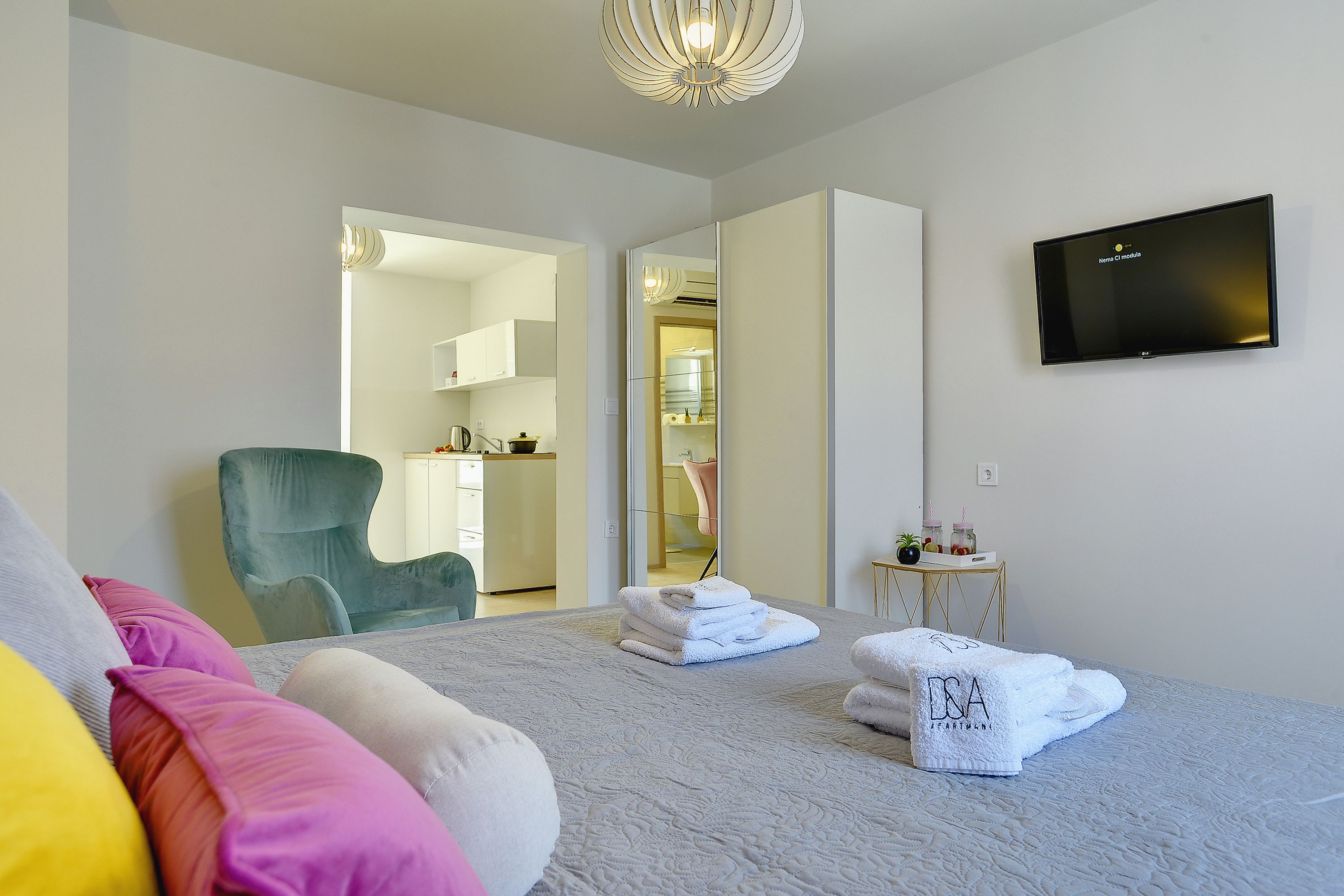 A bedroom with a bed, pillows, a TV, a chair, and a small table with towels.
