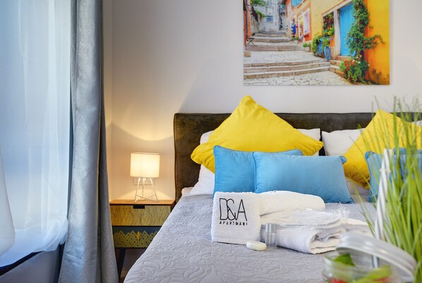 A neatly made bed with a D&A Apartments towel, pillows in yellow and blue, and a bedside lamp.