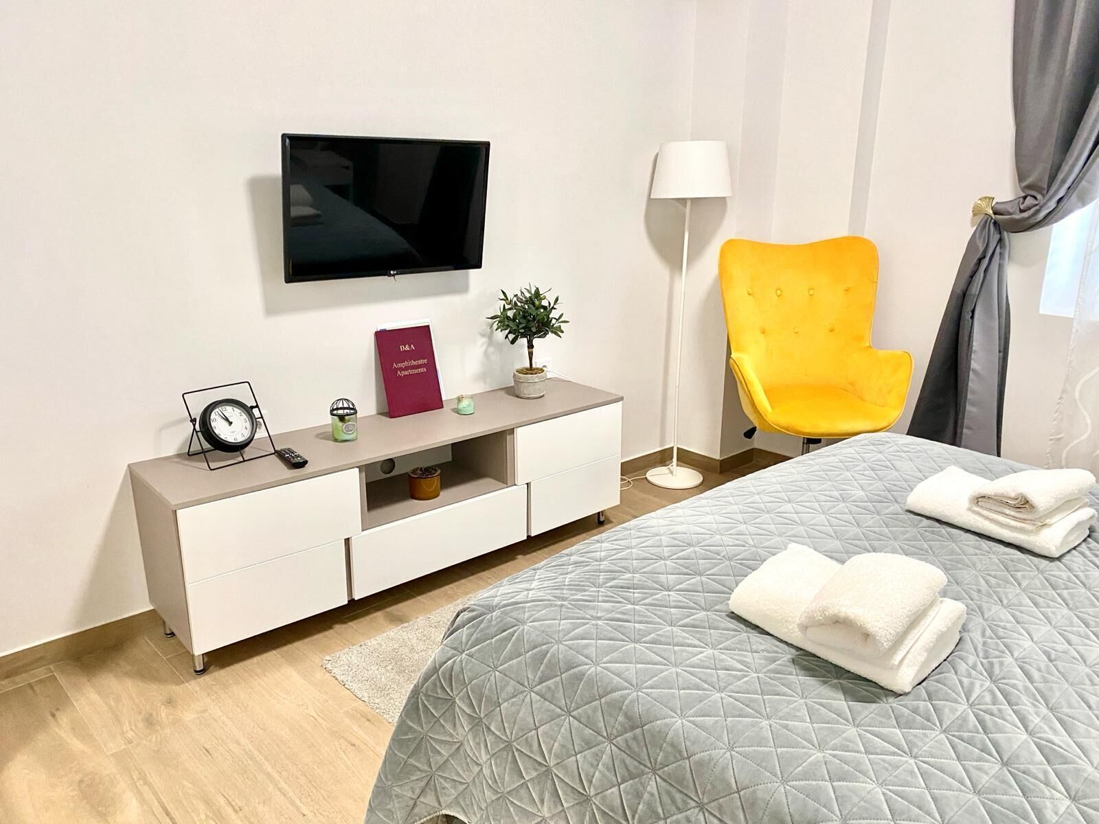 Una habitación de hotel moderna con una cama, un televisor montado en la pared, un sillón amarillo brillante, una pequeña planta en maceta, una lámpara y un letrero con el nombre del hotel.