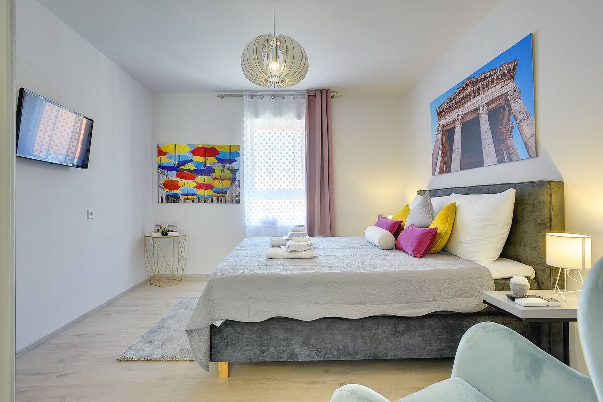 A modern bedroom with a bed, a bedside table, a lamp, a TV, and a painting of a historical building.