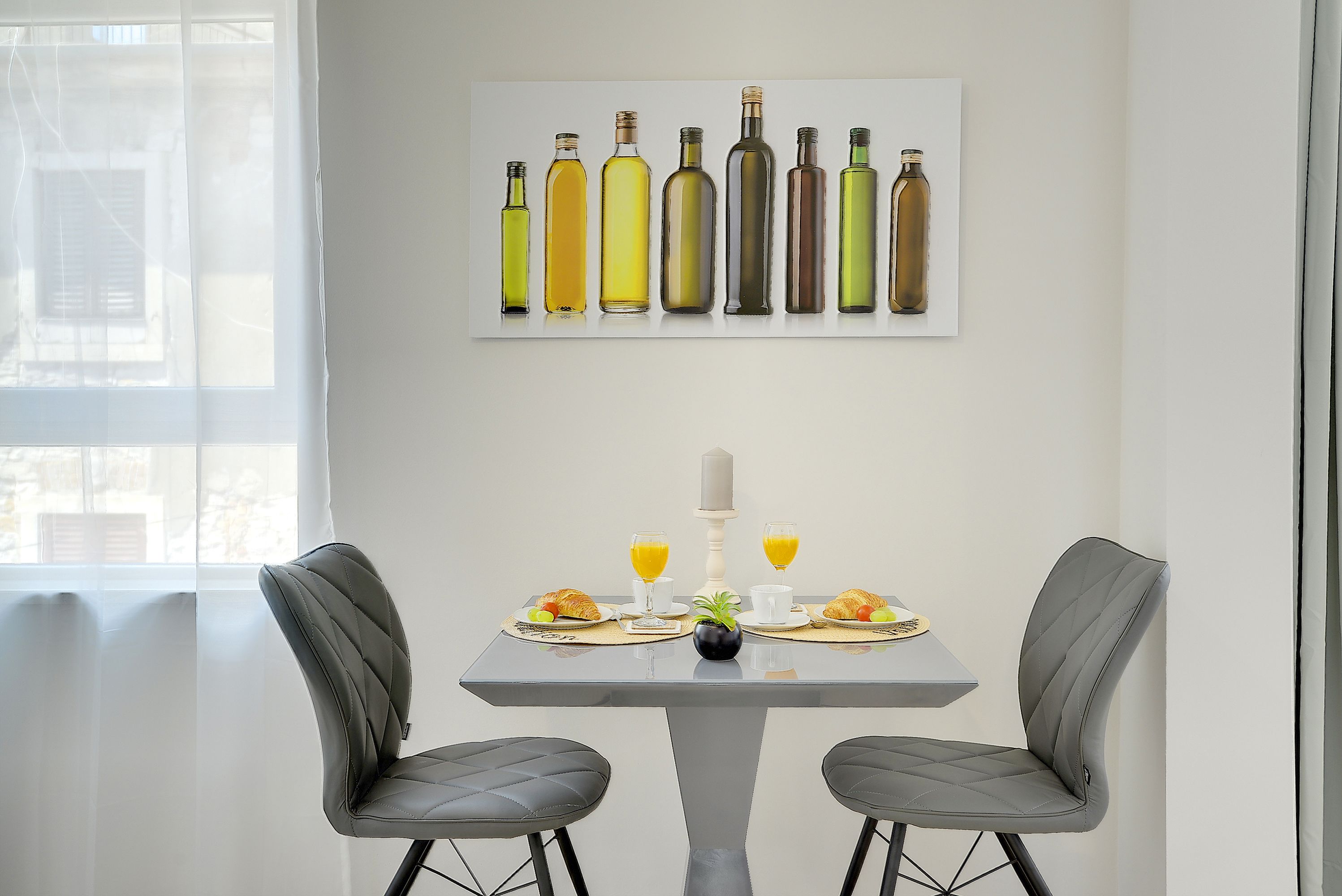 A dining area with a table set for two, featuring a modern chair design, a painting of wine bottles, and a window with curtains.