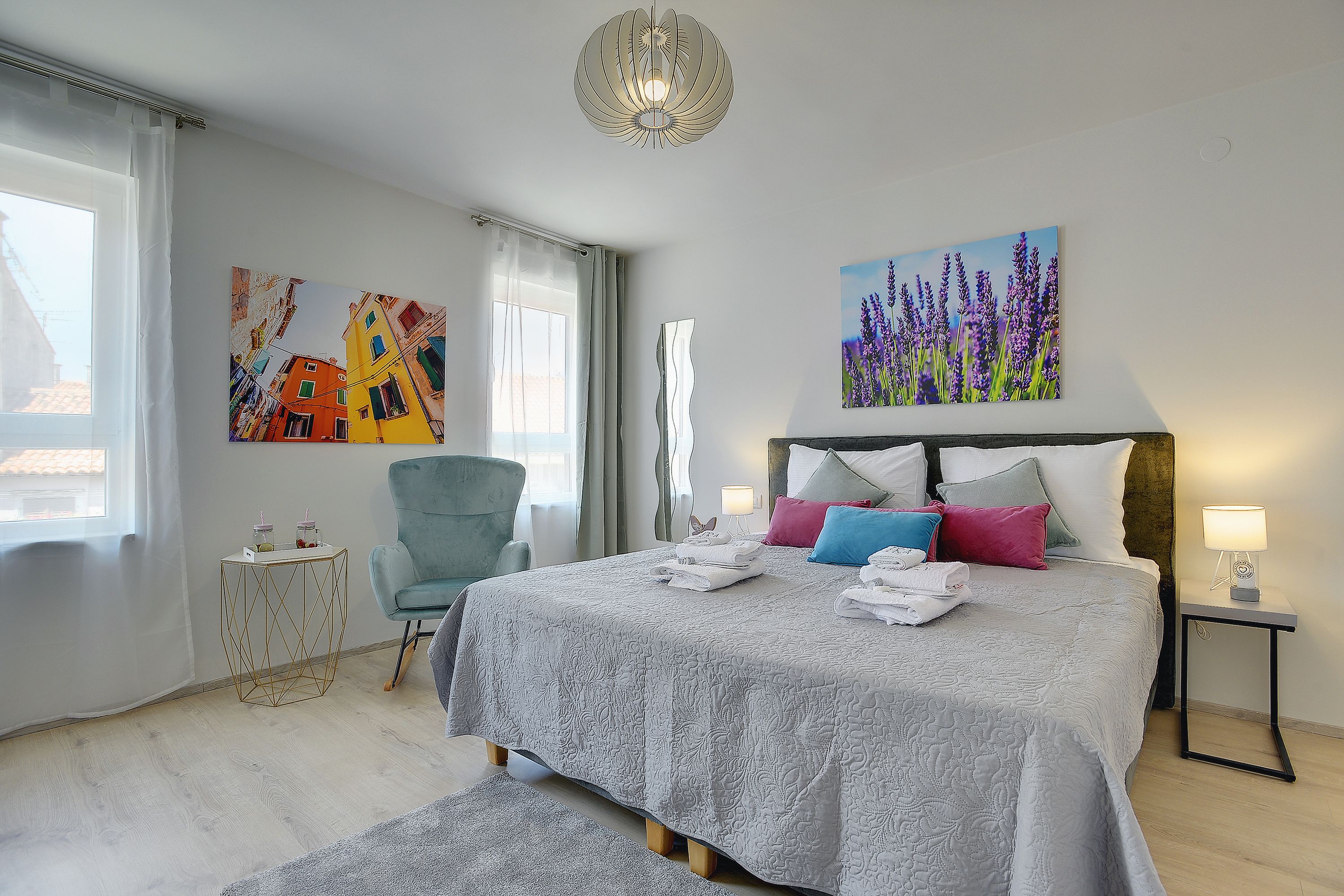 Une chambre à coucher bien rangée, avec un grand lit, une chaise, une table de chevet et deux tableaux accrochés au mur.