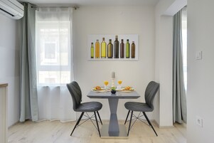 Ein Essbereich mit Tisch für zwei Personen, mit modernem Stuhldesign, einem Gemälde von Weinflaschen an der Wand und einem Fenster mit transparenten Vorhängen.